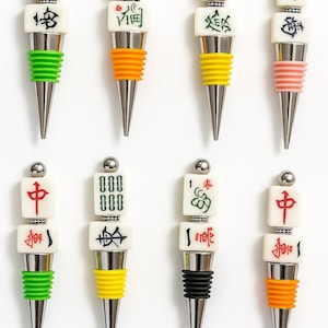 May include: A collection of eight wine bottle stoppers with a silver base and a variety of colorful rubber seals. Each stopper features a different Mahjong tile design, with a silver ball at the top.