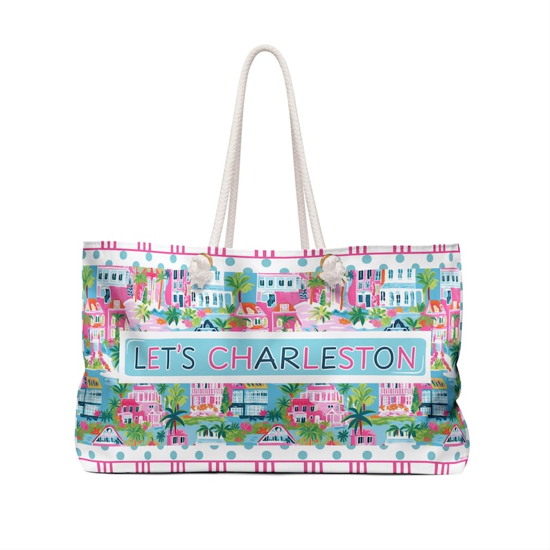 May include: A large tote bag with white rope handles. The bag features a colorful pattern of Charleston houses, palm trees, and the text "LET'S CHARLESTON" on a light blue background. The bag has a white and pink polka dot border.