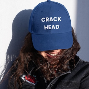 May include: A navy blue baseball cap with the words "CRACK HEAD" in white block letters. The cap is worn by a person wearing a black denim jacket.