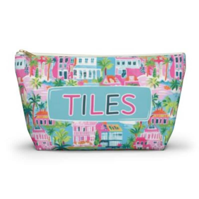 May include: A zippered cosmetic bag with a colorful tropical print featuring pink and white buildings, palm trees, and a blue sky. The word "TILES" is displayed in a blue rectangle.