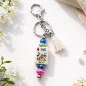 May include: A silver keychain with a white tassel and a charm featuring a tile with a frog design and the text "4 WIN". The charm is adorned with colorful beads in shades of blue, pink, and green.