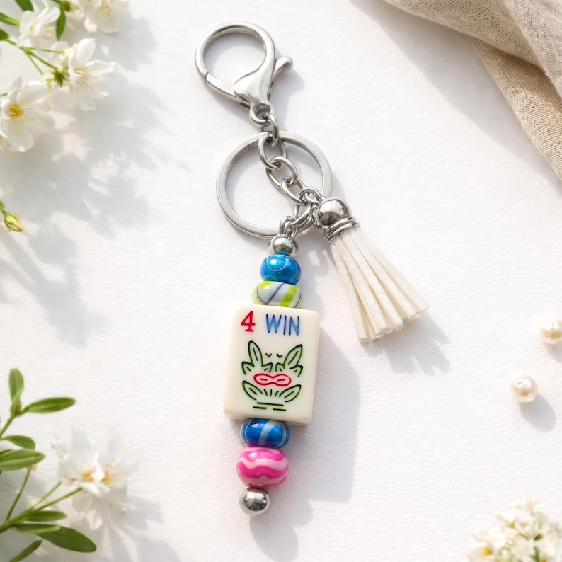May include: A silver keychain with a white tassel and a charm featuring a tile with a frog design and the text "4 WIN". The charm is adorned with colorful beads in shades of blue, pink, and green.