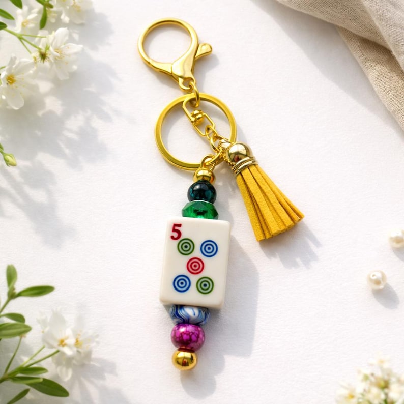 May include: A gold-colored keychain with a white Mahjong tile featuring the number 5 and colorful circles. The keychain includes a yellow tassel, gold beads, and a lobster clasp.