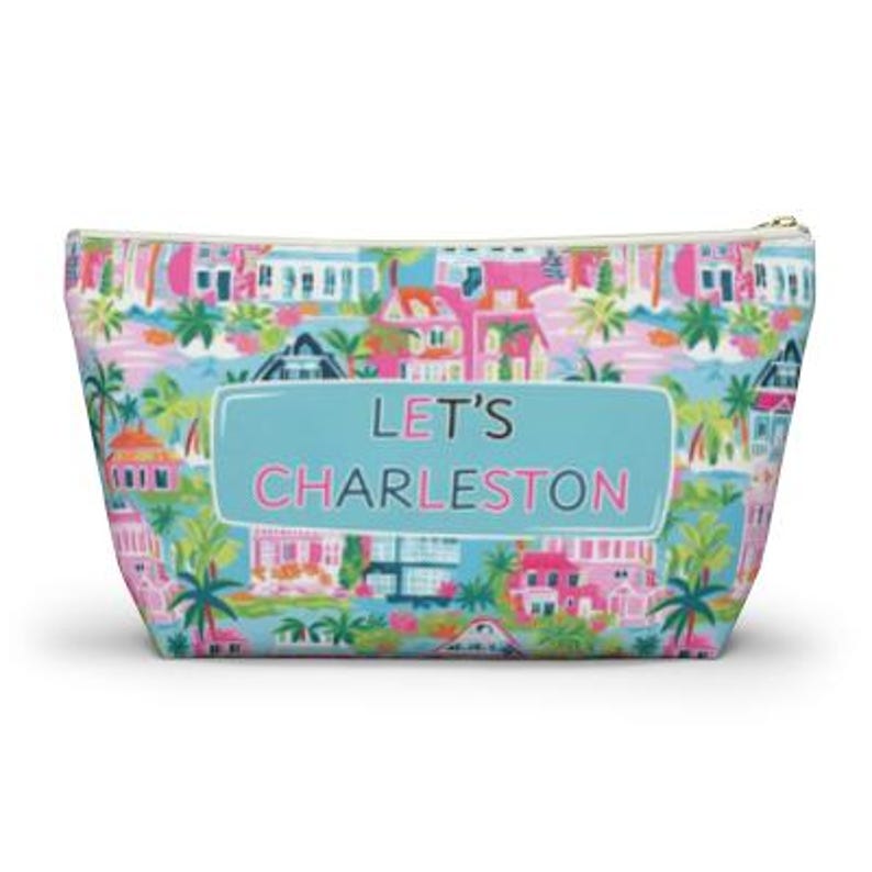 May include: A zippered cosmetic bag with a colorful Charleston-themed pattern. The design features houses, palm trees, and the text "LET'S CHARLESTON" on a light blue background. The bag has a gold zipper.