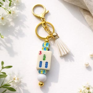 May include: A gold-colored keychain with a white tassel and a charm featuring a Mahjong tile. The tile displays the number 3 and green and blue symbols. The keychain has a gold clasp and a decorative bead design.