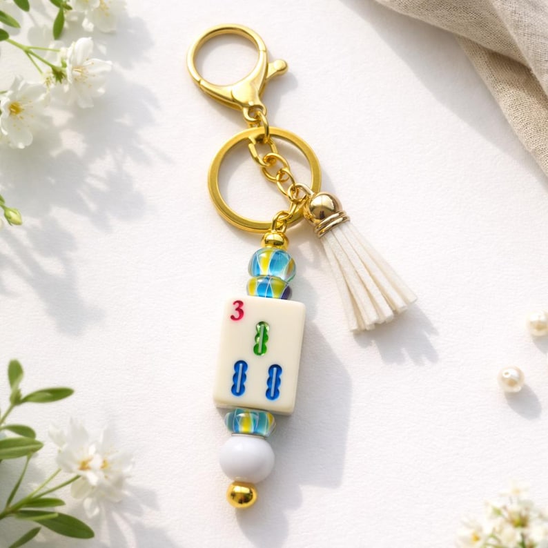 May include: A gold-colored keychain with a white tassel and a charm featuring a Mahjong tile. The tile displays the number 3 and green and blue symbols. The keychain has a gold clasp and a decorative bead design.