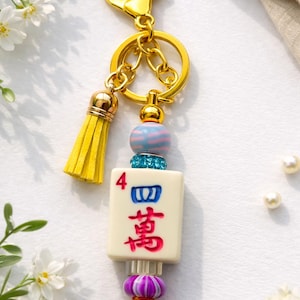 May include: A gold-colored keychain with a yellow tassel and a decorative charm. The charm features a Mahjong tile with red and blue characters, accented with blue and pink beads. The keychain is a decorative accessory.