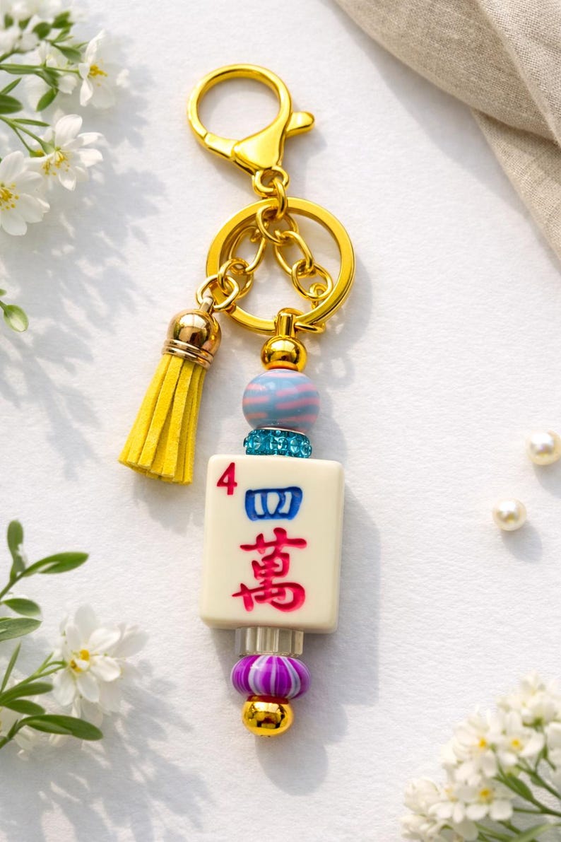 May include: A gold-colored keychain with a yellow tassel and a decorative charm. The charm features a Mahjong tile with red and blue characters, accented with blue and pink beads. The keychain is a decorative accessory.