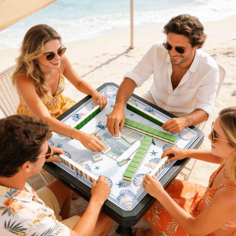 May include: A square Mahjong table with a blue and white ocean-themed design, surrounded by four people playing the game. The table has green tile holders and the word "Mahjong" is visible on a card. The players are on a beach.