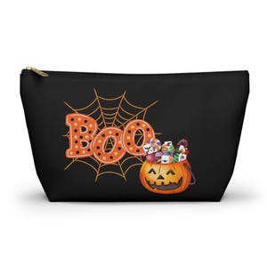 May include: Black zippered cosmetic bag with a Halloween theme. The bag features the word "Boo" in orange, a spiderweb, and a jack-o'-lantern filled with candy. The bag is designed for carrying makeup or other small items.