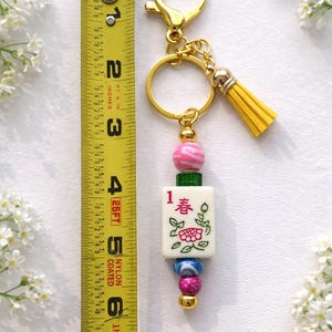 May include: A gold-toned keychain with a yellow tassel and a charm featuring a Mahjong tile. The charm is adorned with pink, green, and blue beads. The tile has a floral design and the number 1 with Chinese characters. The keychain measures approximately 6 inches.