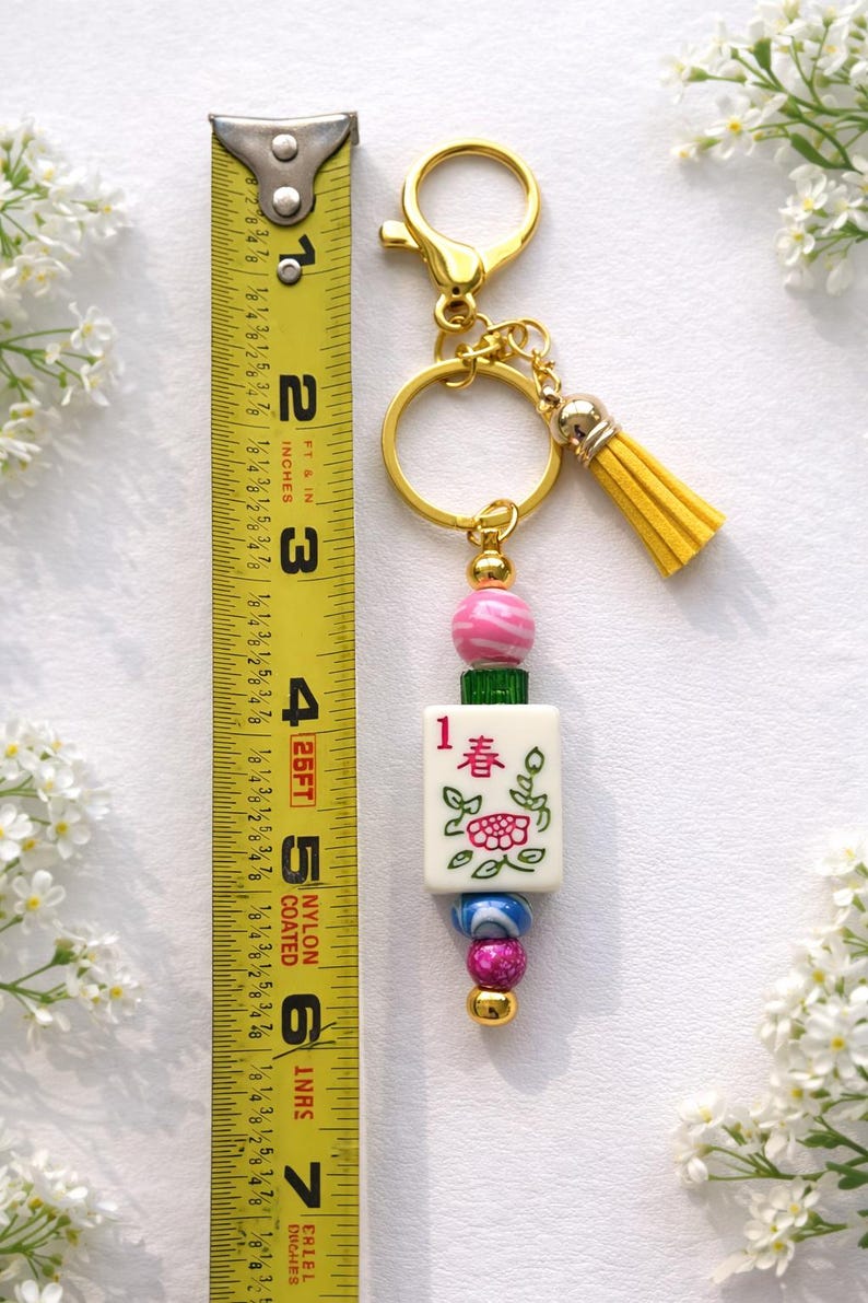 May include: A gold-toned keychain with a yellow tassel and a charm featuring a Mahjong tile. The charm is adorned with pink, green, and blue beads. The tile has a floral design and the number 1 with Chinese characters. The keychain measures approximately 6 inches.