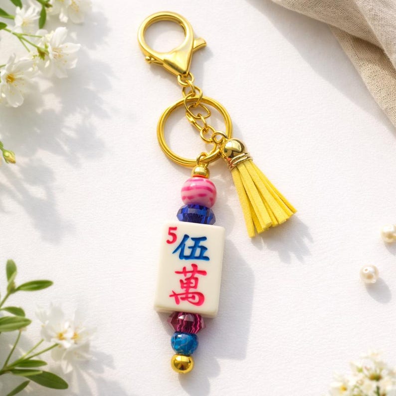 May include: A gold-colored keychain with a white tile featuring red and blue characters, accented with pink, blue, and gold beads, and a yellow tassel. The keychain is designed with a lobster clasp for easy attachment.