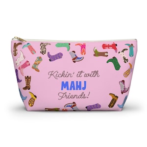 May include: A pink zippered pouch with a pattern of colorful cowboy boots. The text "Kickin' it with MAHJ Friends!" is printed on the front. The pouch has a gold-colored zipper.