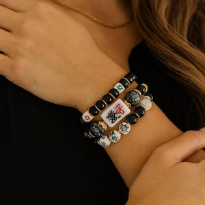 May include: A stack of three bracelets with black, white, and gray beads. One bracelet features a white tile with black Chinese characters. The bracelets are worn on a wrist, with a gold chain visible in the background.
