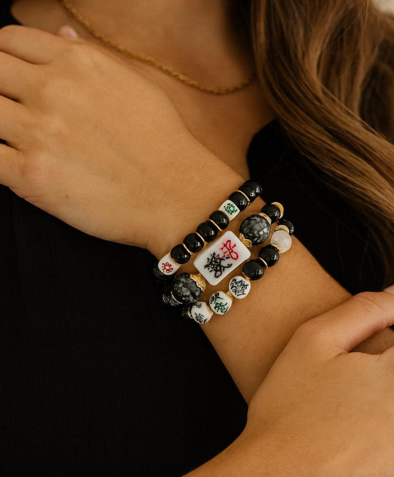 May include: A stack of three bracelets with black, white, and gray beads. One bracelet features a white tile with black Chinese characters. The bracelets are worn on a wrist, with a gold chain visible in the background.