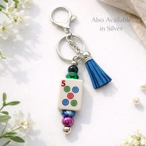 May include: A silver keychain with a blue tassel and a decorative charm. The charm features a white tile with the number 5 and colorful circles, along with a collection of beads in various colors. The text "Also Available in Silver" is visible.