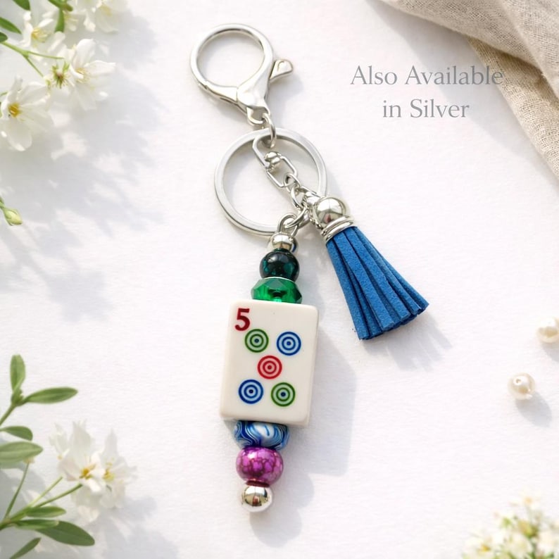 May include: A silver keychain with a blue tassel and a decorative charm. The charm features a white tile with the number 5 and colorful circles, along with a collection of beads in various colors. The text "Also Available in Silver" is visible.