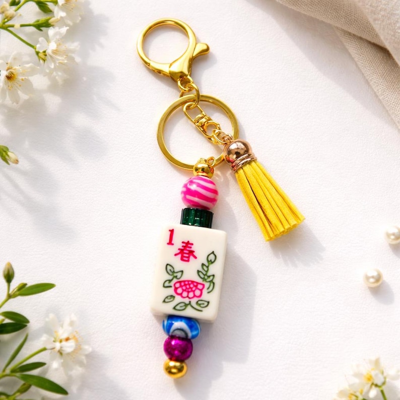May include: A gold-colored keychain with a yellow tassel and a decorative charm. The charm features a white tile with a floral design and the number 1 in red, along with pink and blue beads.