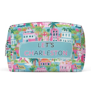 May include: A rectangular cosmetic bag with a colorful Charleston design. The bag features a light blue background with illustrations of buildings, palm trees, and flowers in pink, green, and white. The text "LET'S CHARLESTON" is displayed on the front.