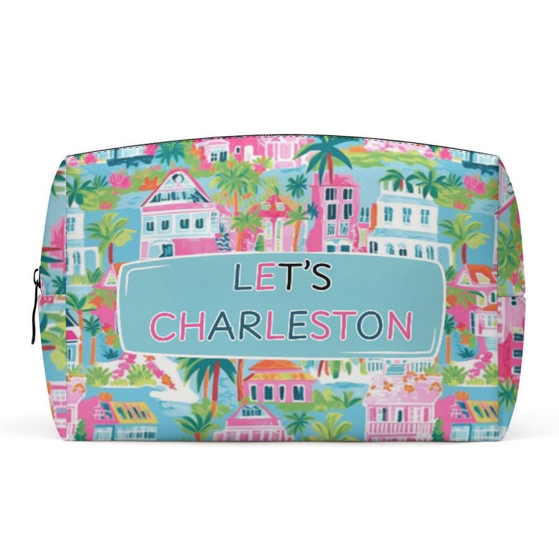 May include: A rectangular cosmetic bag with a colorful Charleston design. The bag features a light blue background with illustrations of buildings, palm trees, and flowers in pink, green, and white. The text "LET'S CHARLESTON" is displayed on the front.