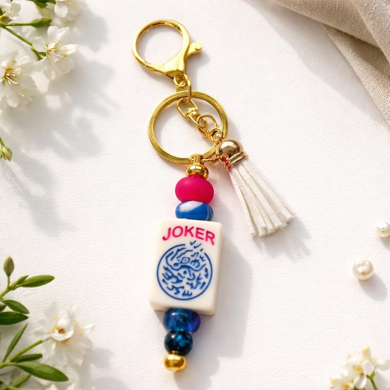 May include: A gold-colored keychain with a white tassel and a charm featuring a tile that says "JOKER". The tile has a blue and white design. The keychain also has pink, blue, and gold beads.