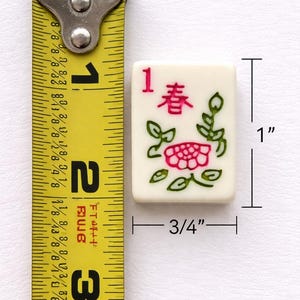 May include: A cream-colored Mahjong tile, measuring 1 inch by 0.75 inches, features the number 1 and Chinese characters in red. A pink flower and green leaves are also depicted. The tile is next to a yellow measuring tape.