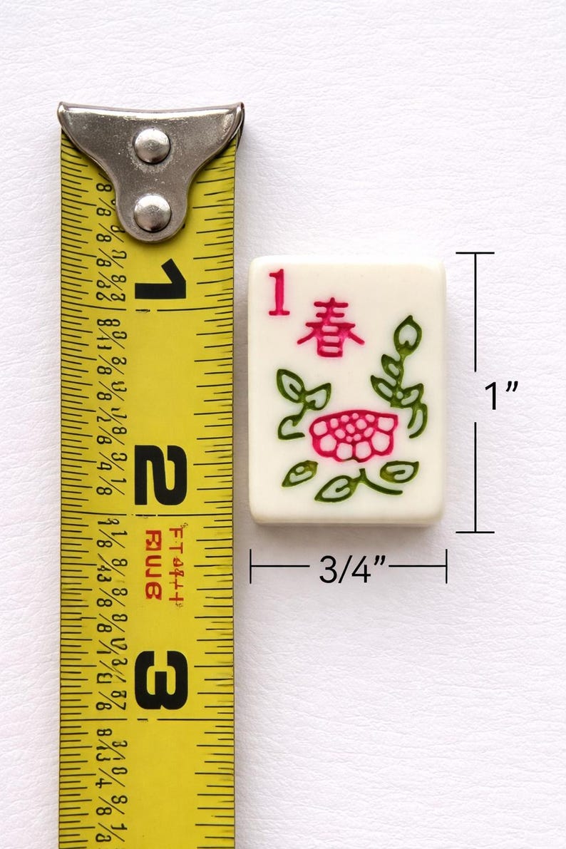 May include: A cream-colored Mahjong tile, measuring 1 inch by 0.75 inches, features the number 1 and Chinese characters in red. A pink flower and green leaves are also depicted. The tile is next to a yellow measuring tape.