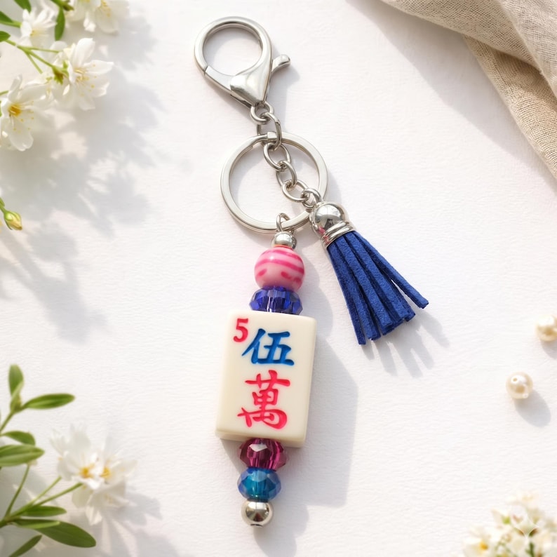 May include: A keychain featuring a white tile with red and blue Chinese characters, accented with pink and blue beads and a navy blue tassel. The keychain has a silver clasp and ring.