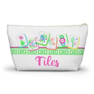 Könnte beinhalten: Weiße Reißverschlusstasche mit rosa und grünem Bordürendesign. Die Tasche zeigt Aquarellillustrationen von Kacheln mit Blumen- und Vogelmotiven. Das Wort "Tiles" ist in rosa Schrift auf der Vorderseite geschrieben.
