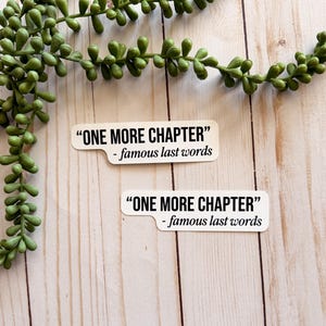 May include: Two white stickers with the text "ONE MORE CHAPTER" and "- famous last words" in black. The stickers are on a light wood grain surface, with a green leafy plant in the upper left corner.
