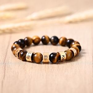 Puede incluir: Una pulsera de cuentas con piedras de ojo de tigre y detalles metálicos dorados. La pulsera incluye cuentas con nombres personalizados. Los nombres "Lily", "Eli" y "Kelly" son visibles. La pulsera se muestra sobre una superficie de madera.