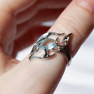 May include: A silver-toned ring featuring a central oval-shaped light blue gemstone. The metal band is designed with intricate, pointed, flame-like or organic swirling cutouts that surround the stone, creating a decorative and detailed setting.