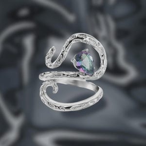 May include: A silver ring with a flowing, ornate design, set with a pear-cut gemstone. The gemstone displays a spectrum of colours, including green, blue, and purple. The ring's band has decorative detailing. A stylish piece of jewellery.