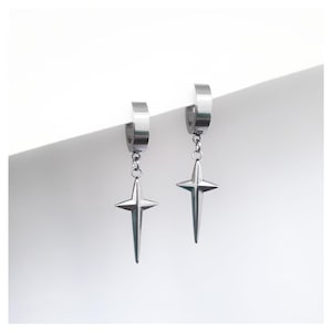 Pode incluir: Um par de brincos de argola prateados com pingentes em forma de estrela. Os brincos têm um acabamento polido e um design simples e moderno. Os pingentes de estrela são simétricos com bordas pontiagudas.