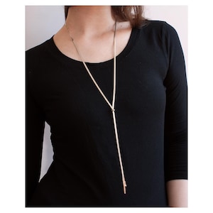 Op de afbeelding: Een lange lariat-ketting in roségoud met een dunne, flexibele ketting die door een kleine, ronde schuifkraal loopt. De ketting hangt in een Y-vorm met twee rechte, slanke metalen staafjes aan de uiteinden, gedragen op een zwart bovenstuk.