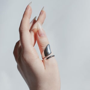 Puede incluir: Un anillo plateado con un diseño geométrico. El anillo presenta una forma triangular que se envuelve alrededor del dedo, con un acabado liso y pulido. El anillo se muestra en una mano con uñas largas y cuidadas.