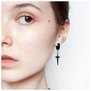 Puede incluir: Un pendiente de aro negro con un colgante de estrella. El pendiente se lleva en la oreja de una persona de piel clara. El colgante de estrella cuelga del aro, creando un diseño simple pero llamativo. El pendiente es de color oscuro.