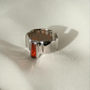 Può includere: Anello argentato con una gemma rettangolare rosso-arancio. L'anello ha un design strutturato e ondulato attorno alla fascia e uno stile aperto e regolabile. La gemma è incastonata al centro dell'anello.