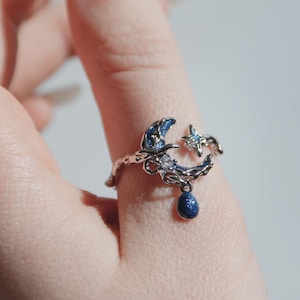 May include: A silver-toned ring featuring a crescent moon, star, and teardrop design. The moon and teardrop are a deep blue with glitter, and the star has a clear gem. The ring is worn on a finger.