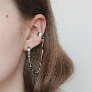 May include: A silver ear cuff and earring set. The ear cuff wraps around the upper ear, connected by a chain to a hoop earring. The jewelry is made of a shiny, metallic material.