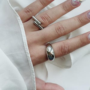 May include: Two silver-toned rings worn on fingers. One ring features a smooth, thick, rounded band with a polished reflective surface. The second ring consists of two parallel, thin, straight metal bars that overlap slightly at the ends.