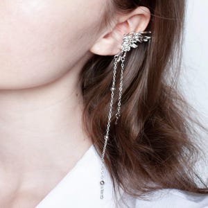 May include: Silver ear cuff featuring a wing design that wraps around the ear. The cuff has dangling chains with small beads and a tiny key charm, creating a delicate and ethereal look. This jewelry piece adds a touch of fantasy to any outfit.