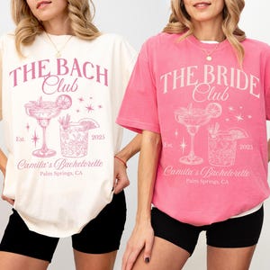 May include: Two white t-shirts with pink text and graphics. The text on the left shirt reads "The Bach Club" and the text on the right shirt reads "The Bride Club". Both shirts feature a cocktail glass with a lime wedge and a starburst design.