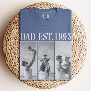 Folded blue unisex t-shirt with ivory “DAD EST. 1995” text and black-and-white three-photo collage showing heartfelt moments between a dad and daughter at the beach. Displayed on a woven round mat in a cozy flat lay style. Perfect custom Father’s Day shirt, birthday gift, or new dad keepsake. Comfort Colors tee with a personal, sentimental touch—ideal for family photo apparel.