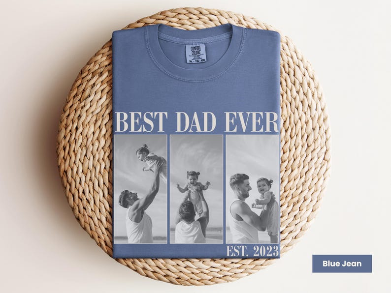 Folded blue jean Comfort Colors t-shirt with bold “BEST DAD EVER” text and a black-and-white three-photo collage of a dad lifting and cuddling his child, with “EST. 2023” at the bottom. Displayed on a woven mat in a cozy flat lay. Personalized photo shirt perfect for new dads, Father’s Day, or baby’s first birthday. A thoughtful and stylish keepsake tee celebrating fatherhood.