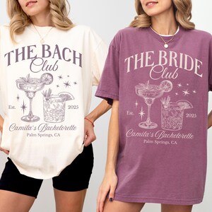 May include: Two oversized t-shirts with a bachelorette party theme. The ivory shirt says "The Bach Club" and the berry shirt says "The Bride Club". Both shirts feature a cocktail glass and a glass of ice with the text "Camila's Bachelorette, Palm Springs, CA" and "Est. 2025".