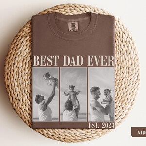 Folded espresso brown Comfort Colors t-shirt with bold “BEST DAD EVER” text and a black-and-white three-photo collage of a father and child, with “EST. 2023” at the bottom. Displayed on a woven mat in a cozy flat lay. Personalized gift shirt for new dads, Father’s Day, or baby’s first birthday. A heartfelt, custom photo tee capturing the love and joy of fatherhood.