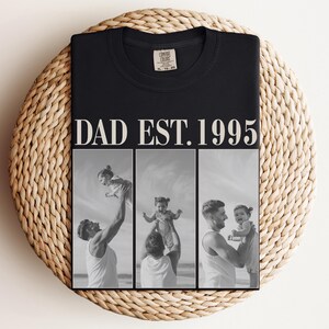 Folded black unisex t-shirt with bold ivory text “DAD EST. 1995” and black-and-white three-photo collage of father and daughter moments at the beach. Displayed on a woven round mat in a flat lay style. Perfect Father’s Day gift, birthday shirt, or new dad keepsake. Soft comfort colors tee with a sentimental, custom design—ideal for personalized dad apparel and photo gifts.