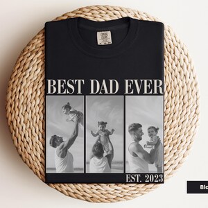 Folded black Comfort Colors t-shirt with bold “BEST DAD EVER” text and a black-and-white three-photo collage of a dad holding and playing with his baby, labeled “EST. 2023.” Displayed on a woven mat in a cozy flat lay. Personalized gift for first-time dads, Father’s Day, or baby’s first birthday. A heartfelt, custom keepsake shirt celebrating the joy of fatherhood.